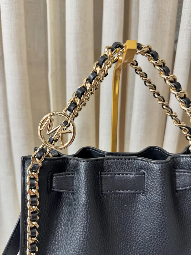 Acercamiento al charm dorado MK y a la cadena entrelazada con cuero negro